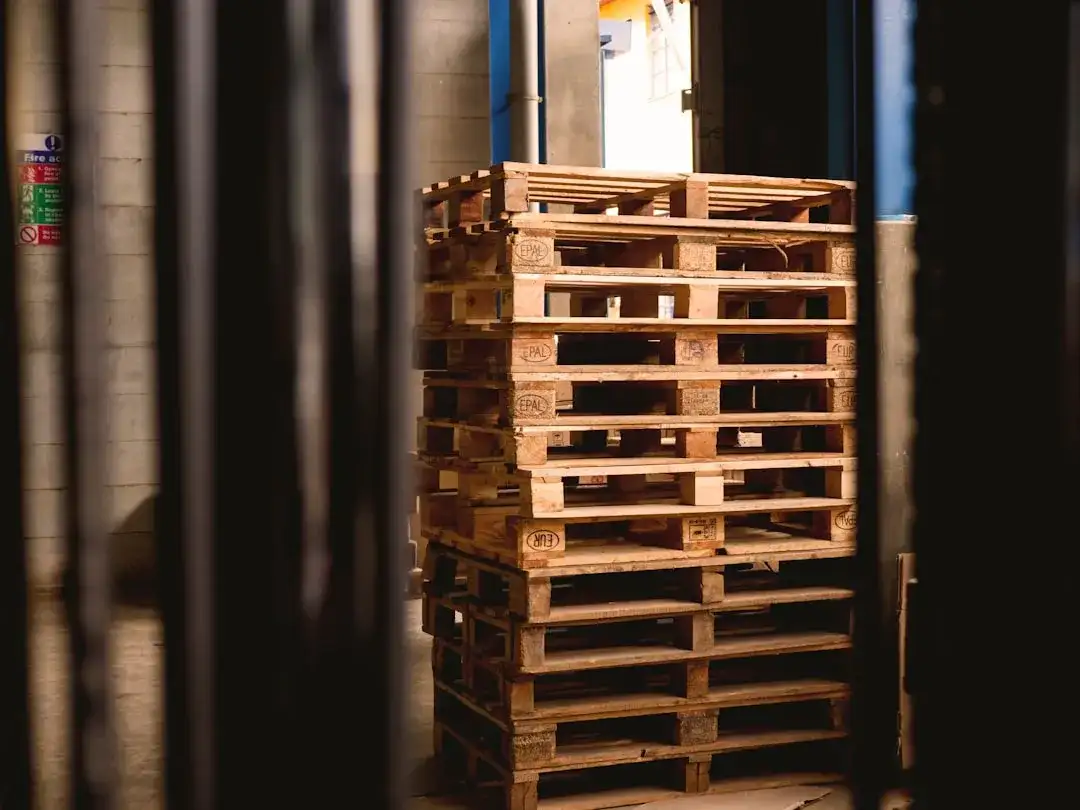 A stack of wooden pallets in a dimly lit warehouse, framed by dark vertical beams in the foreground.